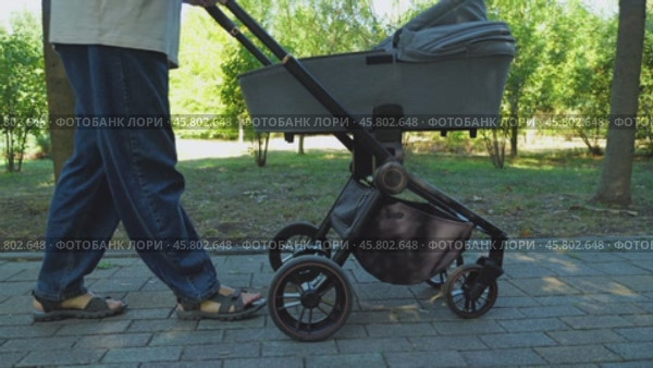 A person in jeans and sandals pushes a black pram with a grey carrycot along a cobbled path in a park. Side view. The camera moves parallel to the pram. Close-up. Стоковое видео, видеограф Воловодов Владимир / Фотобанк Лори