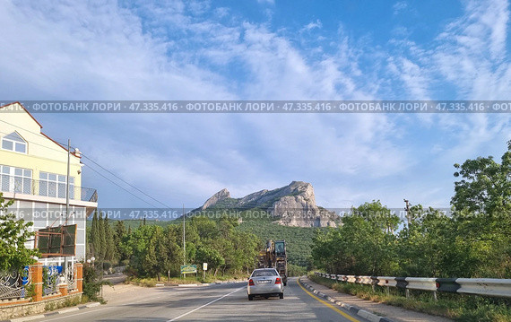 A picturesque corner of Crimea — a road running through green hills at the foot of a mountain, presumably the Kush-Kaya rock massif in the Simeiz region. A passenger car and a tractor with road equipment are moving along the highway. On the left, you can see a well-kept fence with forged elements, a building with an attic, and a billboard offering dishes from the cuisine. On the right and in the distance, there is dense vegetation, and a majestic rock rises above the entire landscape. The clear sky and soft light give the scene a warm, Crimean atmosphere. (2021 год). Редакционное фото, фотограф Анна Кривицкая / Фотобанк Лори