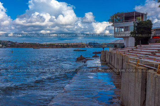A picturesque view of Sevastopol Bay with a concrete embankment and the steps of an amphitheater facing the sea. Along the shore there is a white building with a terrace, and in the distance you can see ships and cranes of shipyards. The weather is sunny but cloudy, creating a striking contrast between the blue sky, sea and architecture. On the horizon you can see the cityscape of Sevastopol with its historical and industrial sites. Стоковое фото, фотограф Анна Кривицкая / Фотобанк Лори