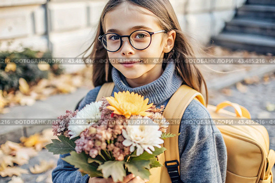 A pretty little girl with glasses and a bouquet of autumn flowers goes to school for the first time. A child with a school bag, a book and flowers. A child on a city street. Back to school. Стоковое AI-изображение, AI-художник Photojuli / Фотобанк Лори
