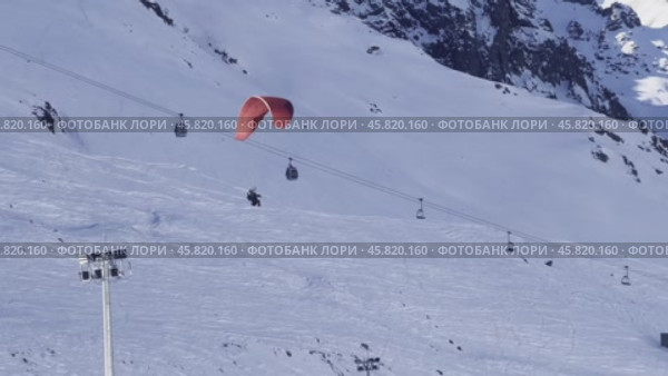 A red paraglider with tourists gently sails through a snow-covered gorge past a mountain slope with a ski slope and a cable car. Редакционное видео, видеограф Ярославский Максим / Фотобанк Лори