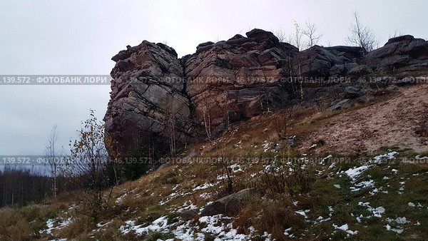 A rock with a characteristic layered structure surrounded by autumn nature. In the foreground, bushes with yellowed leaves are visible, as well as islands of snow on the ground, indicating late autumn or early winter. The rock has a rough texture, many cracks and stratifications, which makes it a unique geological formation.The landscape is typical for the Perm region, where the Ural mountain ranges are combined with forests and open landscapes. Possible location - Vetlan Rock, Chertovo Gorodishche or Stone City. Стоковое фото, фотограф Анна Кривицкая / Фотобанк Лори