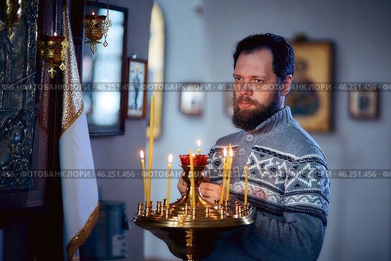 A Russian man with a beard stands in an Orthodox Church, lights a candle and prays in front of the icon. Стоковое фото, фотограф Арзюков Вадим Николаевич / Фотобанк Лори