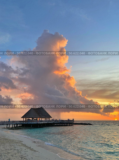 A scenic view of a pier with a thatched-roof structure at sunset, with dramatic clouds and ocean. Concept of: Tropical paradise. Мальдивы (2022 год). Редакционное фото, фотограф Ольга Петрова-Апостолова / Фотобанк Лори