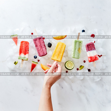 A set of different frozen berry smoothies lolly on a stick with years and fruits. The girl’s hand takes a smoothie on a stick from ice cubes. Flat lay. A woman’s hand holds a frozen fruit smoothie on a stick against the background of ice cubes with berries and fruits. Flat lay. Стоковое фото, агентство Ingram Publishing / Фотобанк Лори