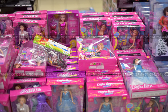 A shop window with bright pink packaging for dolls from various brands, including Abbie, Calleigh and Defa Lucy. The toys are presented in various outfits - evening dresses, ball gowns, accessories. In the center is a set of miniature horses in a transparent bag. The dolls are in neat boxes with a transparent window, allowing you to see their appearance. In the background are store shelves with other toys, creating an atmosphere of children's assortment. (2015 год). Редакционное фото, фотограф Анна Кривицкая / Фотобанк Лори