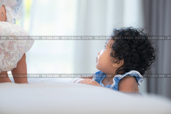 A small girl stood at the foot of the bed playing with the younger sibling carried by his mother and smiling with affection, In a bedroom surrounded by warmth and family. Стоковое фото, агентство Ingram Publishing / Фотобанк Лори