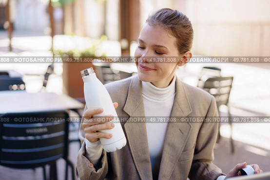 A smiling woman sitting at a caf table, admiring her stylish white water bottle in a vibrant outdoor setting. A Woman Happily Enjoying Her Colorful Water Bottle While Seated in a Cafeteria Setting. Стоковое фото, агентство Ingram Publishing / Фотобанк Лори