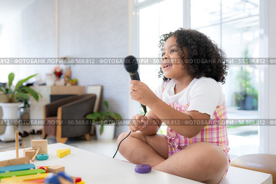 A tiny girl sits on a toy table, clutching a microphone, and sings cheerfully in the living room. Стоковое фото, агентство Ingram Publishing / Фотобанк Лори