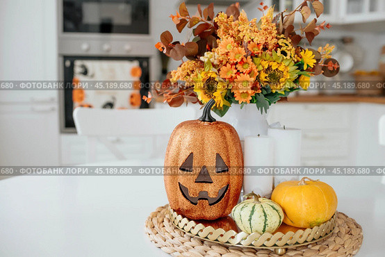 A vase of flowers,a jack pumpkin and candles on a tray. In the background - the interior of a white kitchen in Scandi style. The concept of home and comfort. Autumn decor for the Halloween holiday. Стоковое фото, фотограф Photojuli / Фотобанк Лори