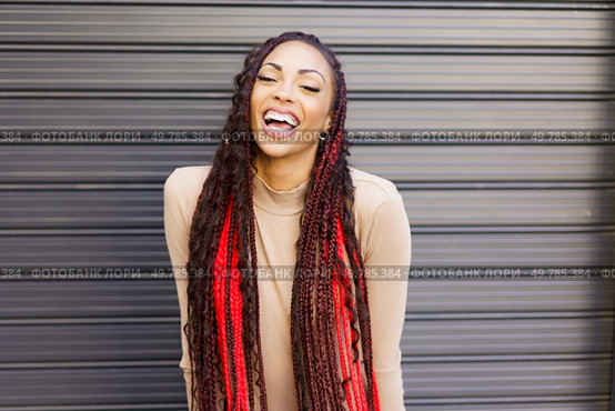 A vibrant and energetic young woman with long, beautifully braided hair adorned with eyecatching red highlights, smiles joyfully and expressively against a richly textured and colorful background. A Joyful Woman with Beautiful Braided Hair Poses Against a Bright Modern Background. Стоковое фото, агентство Ingram Publishing / Фотобанк Лори