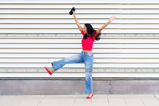 A vibrant young woman, sporting a stylish red top paired with denim jeans, joyfully poses against a creatively striped wall, capturing the true essence of happiness and the spirit of carefree living. A Joyful Young Woman Happily Dancing in a Lively Urban Setting Full of Energy and Life. Стоковое фото, агентство Ingram Publishing / Фотобанк Лори