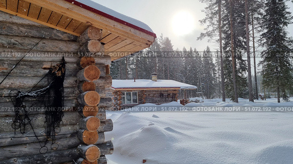 A winter rural landscape with wooden log cabins surrounded by a snow-covered forest. In the foreground is the corner of a log house with a decorative net leaning against the wall. In the distance, another log house is visible with a roof covered with a thick layer of snow and a chimney from which smoke may be coming. On the facade of the second house hangs a sign with the inscription "Teremok". Sunlight breaks through the light snowfall, creating an atmosphere of coziness and silence. Tall pines and firs emphasize the beauty of northern nature, and untouched snow adds a feeling of freshness and peace. Стоковое фото, фотограф Анна Кривицкая / Фотобанк Лори
