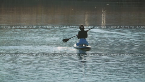 A woman floating on a paddleboard on a calm body of water reflecting the sun's glare. She is wearing a black top and jeans. Back view. Riding on sup board. Стоковое видео, видеограф Воловодов Владимир / Фотобанк Лори