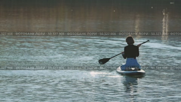 A woman sitting on her knees on a paddleboard paddling on a calm lake. She has a magnificent figure. The soft ripples reflect the sunlight. Стоковое видео, видеограф Воловодов Владимир / Фотобанк Лори