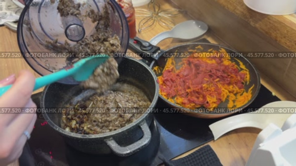A woman transfers chopped mushrooms from a blender bowl to a saucepan with fried onions. Nearby, in a frying pan, is fried carrot with tomato paste. She is preparing mushroom caviar. Стоковое видео, видеограф Vadzim Yakubovich / Фотобанк Лори