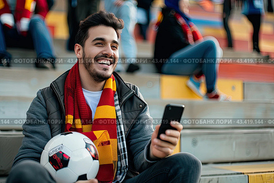A young man is sitting in the stadium wearing a branded scarf and holding a smartphone and a soccer ball in his hands. Стоковое AI-изображение, AI-художник Ярославский Максим / Фотобанк Лори