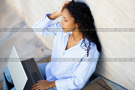 A young woman dressed in a blue striped shirt is deeply immersed in her thoughts, diligently working on her laptop while casually seated against a wall in a pleasant outdoor environment. A woman in blue stripes works on her laptop outdoors, combining leisure with productivity. Стоковое фото, агентство Ingram Publishing / Фотобанк Лори