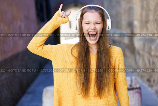 A young woman, dressed in a bright yellow sweater, wearing stylish headphones, joyfully expresses herself as she immerses in the music, fully embracing the vibrant urban atmosphere all around her. A Joyful Young Woman Dances While Listening to Music in a Bright and Vibrant Alleyway. Стоковое фото, агентство Ingram Publishing / Фотобанк Лори
