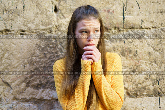 A young woman, dressed in a cozy yellow sweater, stands against an old, rustic stone wall, appearing worried. Her expression poignantly captures a moment filled with anxiety or deep contemplation. A Young Woman Dressed in a Yellow Sweater is Expressing Concern Against a Textured Wall. Стоковое фото, агентство Ingram Publishing / Фотобанк Лори