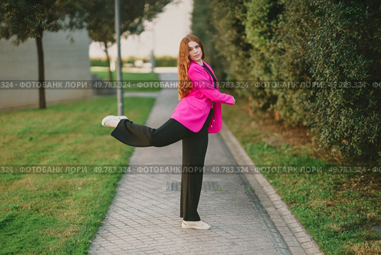 A young woman, elegantly and stylishly dressed in a vibrant pink blazer, poses joyfully in an outdoor setting along a sunlit pathway surrounded by lush greenery, radiating confidence and charm. A Stylish Young Woman Dressed in a Pink Blazer is Posing Outdoors with Great Joy. Стоковое фото, агентство Ingram Publishing / Фотобанк Лори