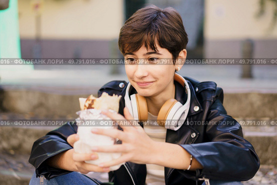 A young woman in a trendy jacket sits on steps in a lively outdoor area, enjoying music through her headphones and a delicious dessert, creating a joyful vibe in a bustling urban setting. A Young Woman Fully Enjoying a Delicious Dessert While Wearing Headphones in an Urban Setting. Стоковое фото, агентство Ingram Publishing / Фотобанк Лори