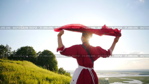 A young woman in russian traditional clothes walking on the green field - her red handkerchief flutters by the wind. Стоковое видео, видеограф Константин Шишкин / Фотобанк Лори