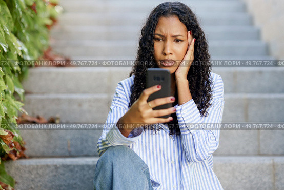 A young woman sits on outdoor steps, shocked and worried, intently checking her phone. Her expressive face shows disbelief and surprise, reflecting her intense feelings in this moment. A worried woman sits on the steps, anxiously looking at her phone amid vibrant city life. Стоковое фото, агентство Ingram Publishing / Фотобанк Лори