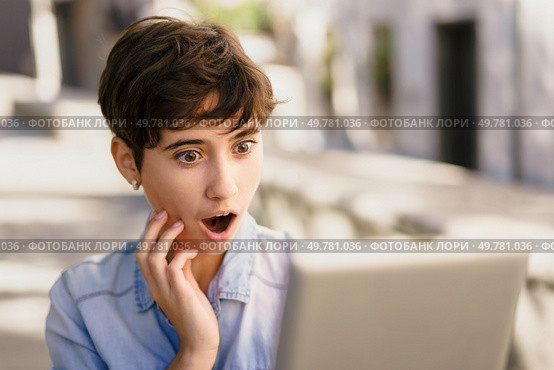 A young woman with a stylish short haircut looks at her laptop outdoors, her face showing surprise and curiosity about an unexpected online discovery, reflecting her engagement with the digital world. A Young Woman Looks Surprised and Reacts While Viewing Her Laptop Screen Outdoors. Стоковое фото, агентство Ingram Publishing / Фотобанк Лори