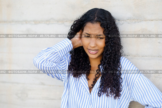 A young woman with beautiful curly hair appears to be deep in thought, as she stands gracefully against a textured concrete wall, wearing a stylish striped shirt that showcases her personality. A Young Woman with Curly Hair, Dressed in a Striped Shirt, Stands Against a Concrete Wall. Стоковое фото, агентство Ingram Publishing / Фотобанк Лори