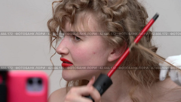A young woman with long hair is using a curling wand to style hair, manually wraps sections of blonde hair around the wand. Broadcast live video to social media (2024 год). Редакционное видео, видеограф А. А. Пирагис / Фотобанк Лори