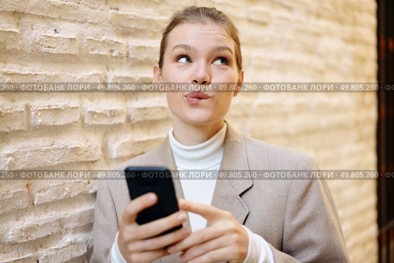 A youthful woman, wearing a contemplative and pensive look on her face, is absorbed in using her smartphone while casually leaning against a rugged and textured brick wall in broad daylight. A thoughtful woman is using her smartphone while standing against a textured brick wall outside. Стоковое фото, агентство Ingram Publishing / Фотобанк Лори