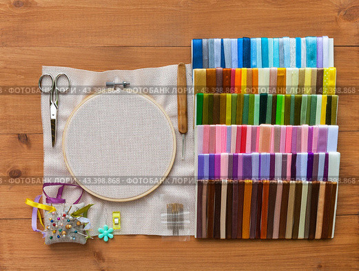 Accessories for hand embroidery with satin ribbons: canvas in hoop, pincushion with needles, set of multi-colored satin ribbons, scissors, hook and needle threader on wooden table. Flat lay, closeup, mockup. Стоковое фото, фотограф Виктория Катьянова / Фотобанк Лори
