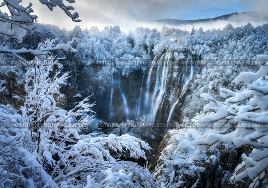 Aerial drone view of amazing waterfall and snowy forest at sunset. Winter fairytale in Plitvice Lakes, Croatia. Top view of white snow-covered frozen trees, waterfall, house, clouds on cold winter day. Стоковое фото, агентство Ingram Publishing / Фотобанк Лори