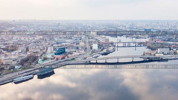 Aerial panoramic view from the drone on the central historical part of the city Kiyv and the district of Podol with Rybalskiy Island, churches, the ships, river docks and Havana bridge. Reflection of sunset and cloudy sky in the Dnieper River.. The panoramic bird’s eye view shooting from drone of the Podol district, the right bank of the Dnieper River and Rybalskiy Island in Kiev, Ukraine at summer sunset on the background of the cloudy sky. Стоковое фото, агентство Ingram Publishing / Фотобанк Лори