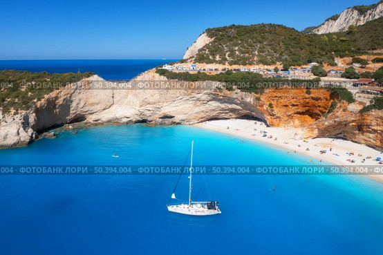 Aerial view of blue sea, yacht, rock, sandy beach with umbrellas at sunny day in summer. Porto Katsiki, Lefkada island, Greece. Beautiful landscape with sea coast, boat, azure water. Top view. Travel. Стоковое фото, агентство Ingram Publishing / Фотобанк Лори