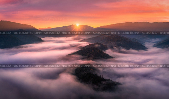Aerial view of mountains in low clouds at sunrise in autumn. Top view of mountain hills in fog in fall at dawn. Beautiful landscape with ridges, forest, sun, colorful orange sky with pink clouds. Стоковое фото, агентство Ingram Publishing / Фотобанк Лори