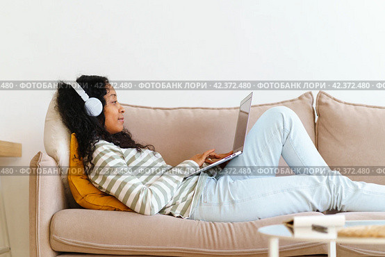 African-American female in headphones surfing the net on laptop while lying on sofa in living room. Стоковое фото, фотограф Natalia Gdovskaia / Фотобанк Лори
