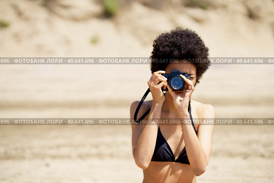 African American female photographer in swimwear taking picture on photo camera while standing on sandy shore during trip in tropical resort. Black woman in bikini taking photo on beach. Стоковое фото, агентство Ingram Publishing / Фотобанк Лори
