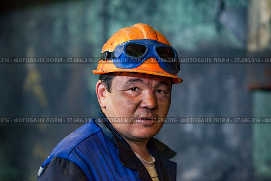 Aksu, Pavlodar region, Kazakhstan - May 29, 2012: Aksu steam power station. Portrait of adult Asian worker in orange hardhat. With welding protective glasses. Close up photo. Blurred background. Редакционное фото, фотограф Alexey Rezvykh / Фотобанк Лори