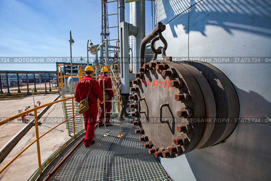 Aktobe region/Kazakhstan - May 04 2012: Oil refinery plant. Two maintenance workers in red work wear and yellow helmets on refining column. On pipelines and blue sky background. CNPC company. Редакционное фото, фотограф Alexey Rezvykh / Фотобанк Лори