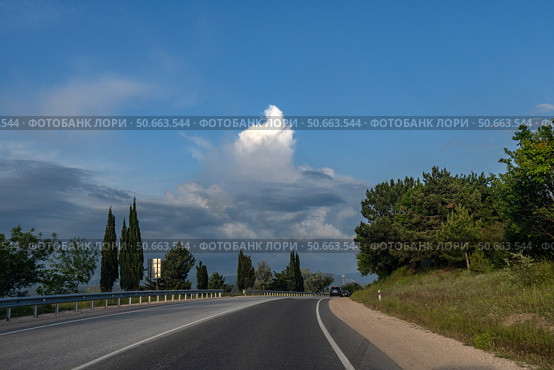 An asphalt road in Crimea, curving among green hills and cypresses against a dramatic sky with voluminous clouds. On the right side of the road there is dense greenery and pine trees, and on the left there is a metal fence and lonely cars. The warm evening light emphasizes the textures of the clouds and creates an atmosphere of peace and an approaching thunderstorm. Стоковое фото, фотограф Анна Кривицкая / Фотобанк Лори