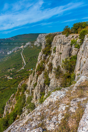 An impressive rocky cliff in the Crimean mountains overlooking a serpentine road that stretches along wooded slopes. In the foreground is a textured cliff surface covered in dry grass and moss. In the distance are mountain ranges, partly covered in greenery, under a bright blue sky with light clouds. Epic and picturesque landscape is perfect for themes of active recreation, mountaineering, hiking and natural power. Стоковое фото, фотограф Анна Кривицкая / Фотобанк Лори