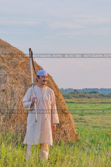 An senior Muslim man in a skullcap and traditional clothes leaves with a hand scythe from the hay field at sunset. Стоковое фото, фотограф Ирина Кононова / Фотобанк Лори
