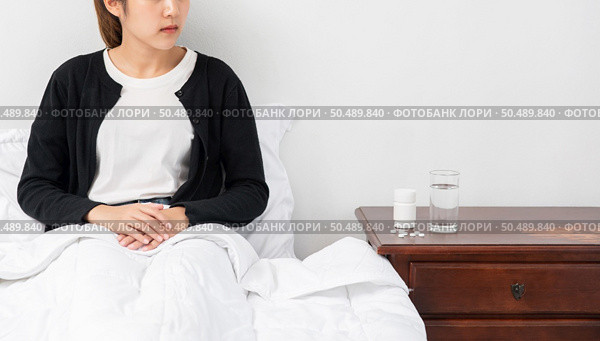 An uncomfortable woman sits on the bed and has medicine on the table. Стоковое фото, агентство Ingram Publishing / Фотобанк Лори