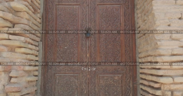 Antique wooden carved door with intricate oriental floral patterns set in sunlit yellow brick wall, historical architecture detail with traditional craftsmanship and aged surface texture from Asia. Редакционное видео, видеограф Сергей Тимофеев / Фотобанк Лори
