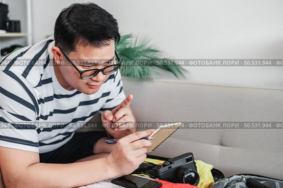 Asian man glasses is preparing clothes in suitcases. He’s choosing clothes, travel documents, the itinerary for a solo trip, and check the checklist. Travel, holiday and vacation concept. Стоковое фото, агентство Ingram Publishing / Фотобанк Лори