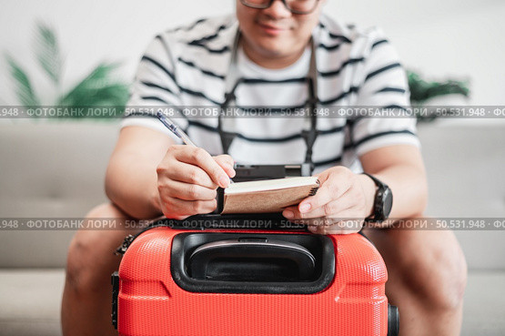 Asian man glasses is preparing clothes in suitcases. He’s choosing clothes, travel documents, the itinerary for a solo trip, and check the checklist. Travel, holiday and vacation concept. Стоковое фото, агентство Ingram Publishing / Фотобанк Лори