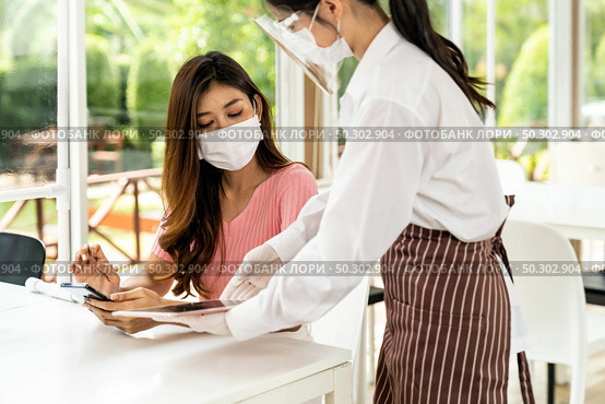 Asian waitress wear facemask and face shield using tablet to show restaurant electronic menu and recommend to customer. New normal technology and lifestyle food and dining concept. Стоковое фото, агентство Ingram Publishing / Фотобанк Лори