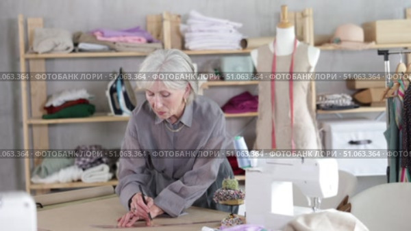 Attentive old tailoress recording dress design on large paper in sewing studio. Стоковое видео, видеограф Яков Филимонов / Фотобанк Лори